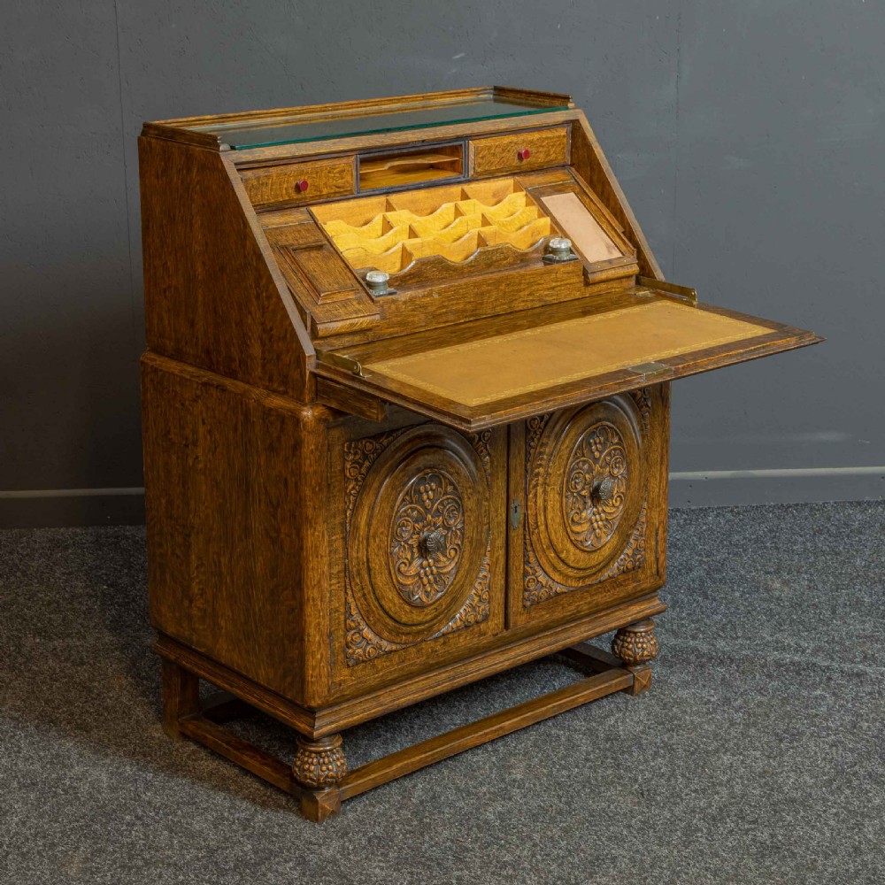 A medium brown carved oak writing bureau with a slant front lid above a double door cupboard, detailed with carved oval foliate panels and turned feet. View XV