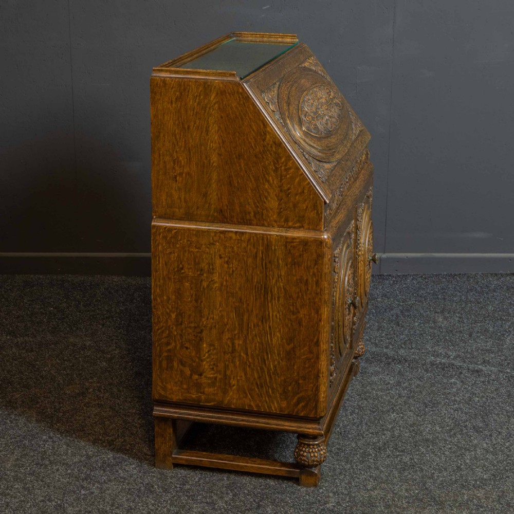 A medium brown carved oak writing bureau with a slant front lid above a double door cupboard, detailed with carved oval foliate panels and turned feet. View XVI