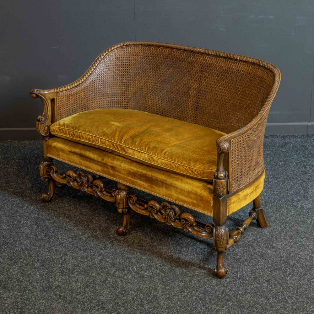 A William and Mary style walnut sofa featuring a high curved woven cane backrest, gold velvet cushion seat, and intricately carved scrollwork base.