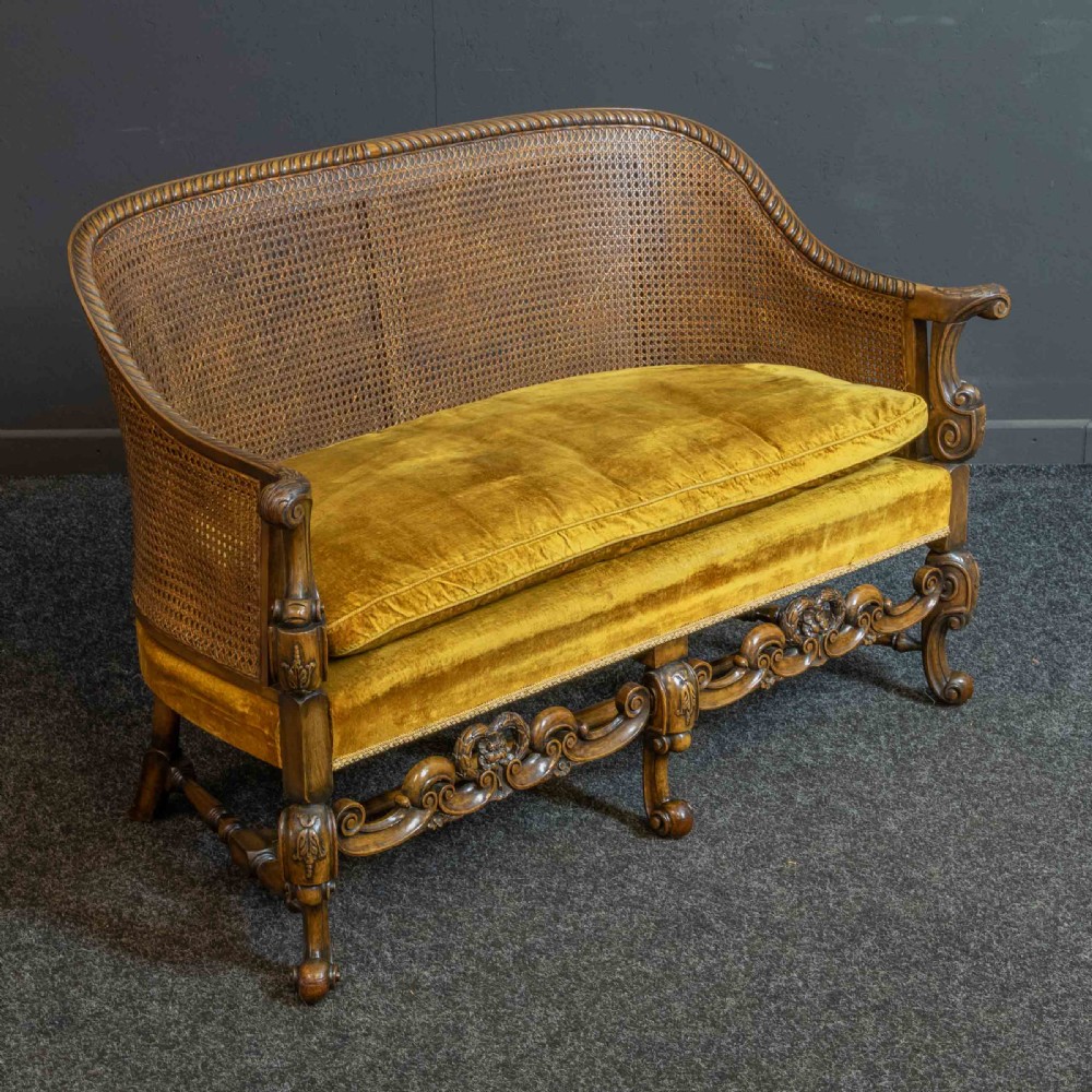A William and Mary style walnut sofa featuring a high curved woven cane backrest, gold velvet cushion seat, and intricately carved scrollwork base. View VI