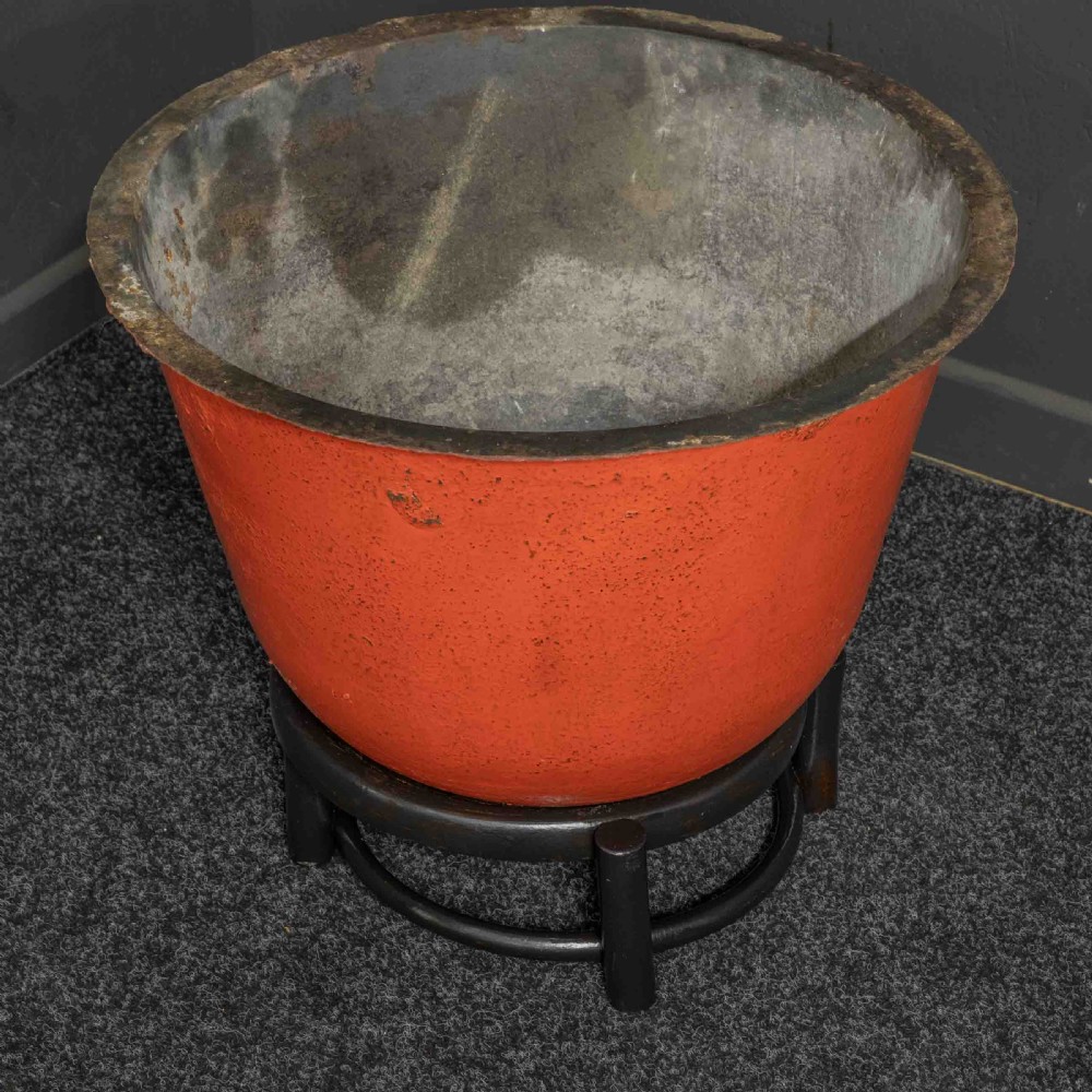 A deep circular copper tub painted orange on the outer side, resting in a black circular stand. View V