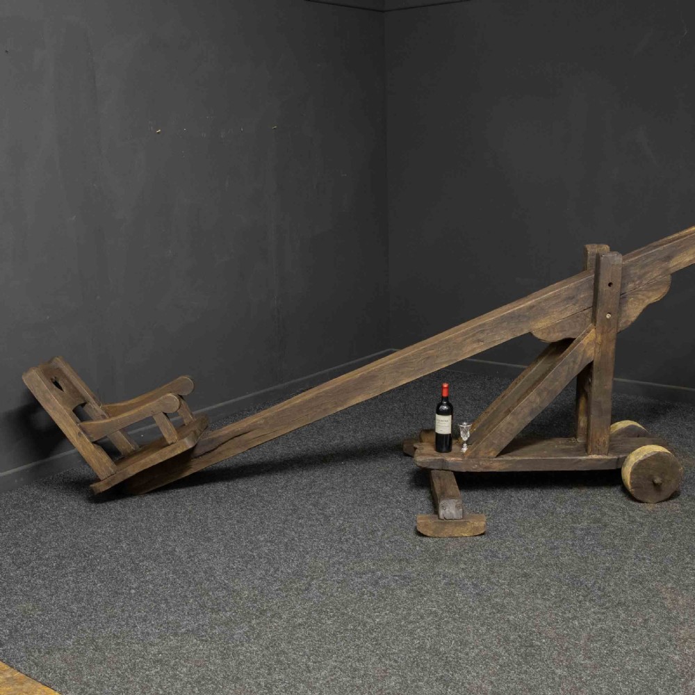 A large dark timber ducking stool featuring a long lever beam, a seated wooden chair at one end, and a heavy support frame on wooden wheels. View II