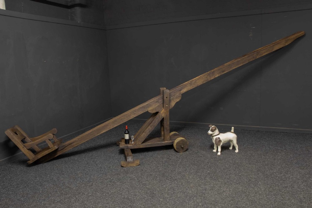 A large dark timber ducking stool featuring a long lever beam, a seated wooden chair at one end, and a heavy support frame on wooden wheels. View III