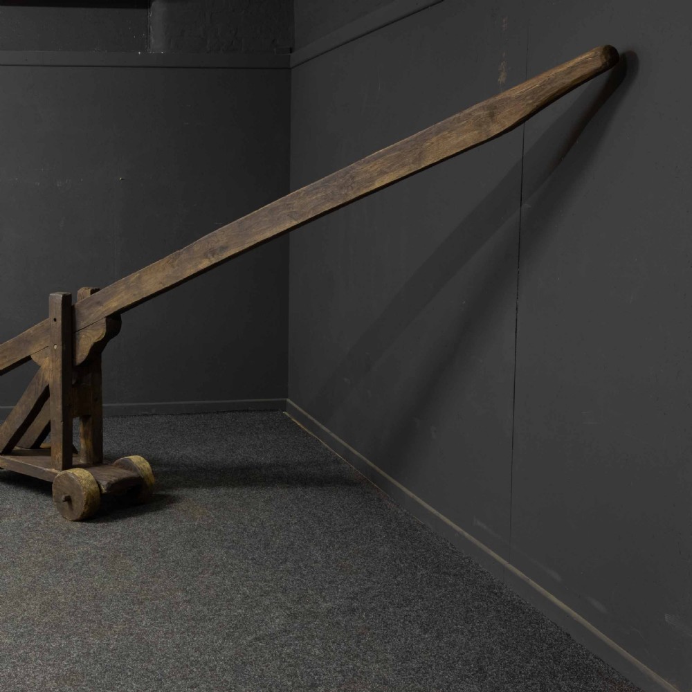 A large dark timber ducking stool featuring a long lever beam, a seated wooden chair at one end, and a heavy support frame on wooden wheels. View IV