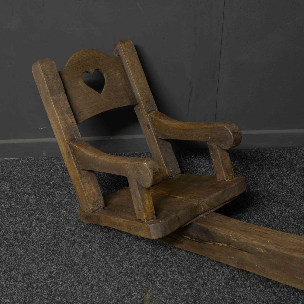 A large dark timber ducking stool featuring a long lever beam, a seated wooden chair at one end, and a heavy support frame on wooden wheels. View V