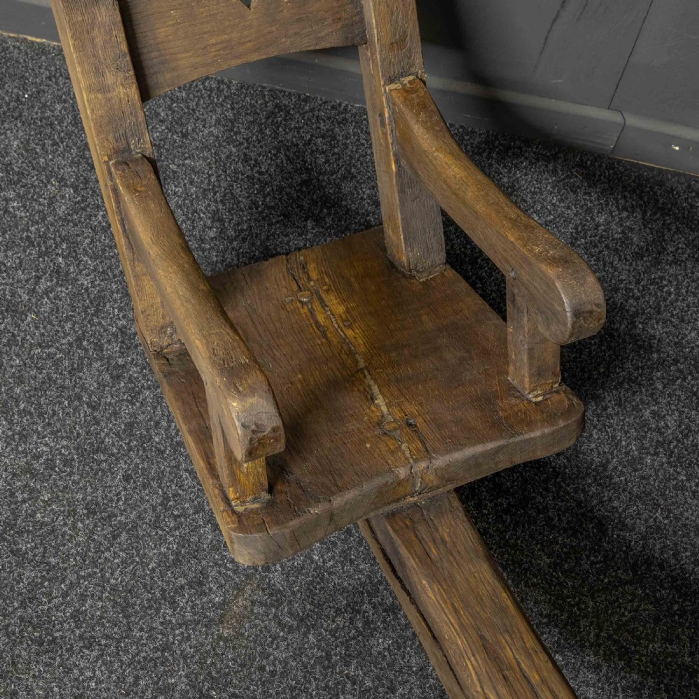A large dark timber ducking stool featuring a long lever beam, a seated wooden chair at one end, and a heavy support frame on wooden wheels. View VI