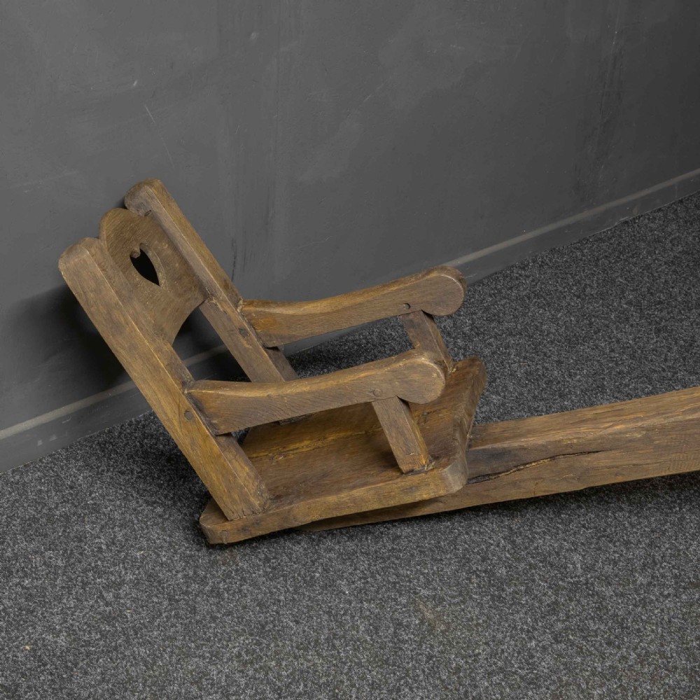A large dark timber ducking stool featuring a long lever beam, a seated wooden chair at one end, and a heavy support frame on wooden wheels. View VIII
