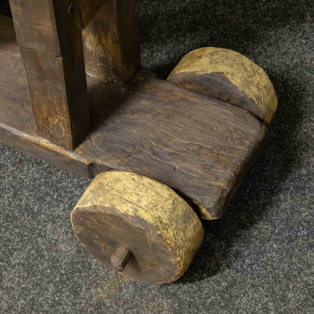 A large dark timber ducking stool featuring a long lever beam, a seated wooden chair at one end, and a heavy support frame on wooden wheels. View XI
