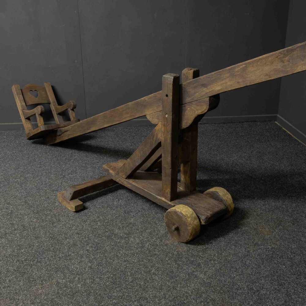 A large dark timber ducking stool featuring a long lever beam, a seated wooden chair at one end, and a heavy support frame on wooden wheels. View XIII