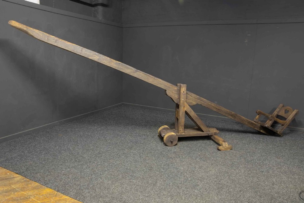 A large dark timber ducking stool featuring a long lever beam, a seated wooden chair at one end, and a heavy support frame on wooden wheels. View XV