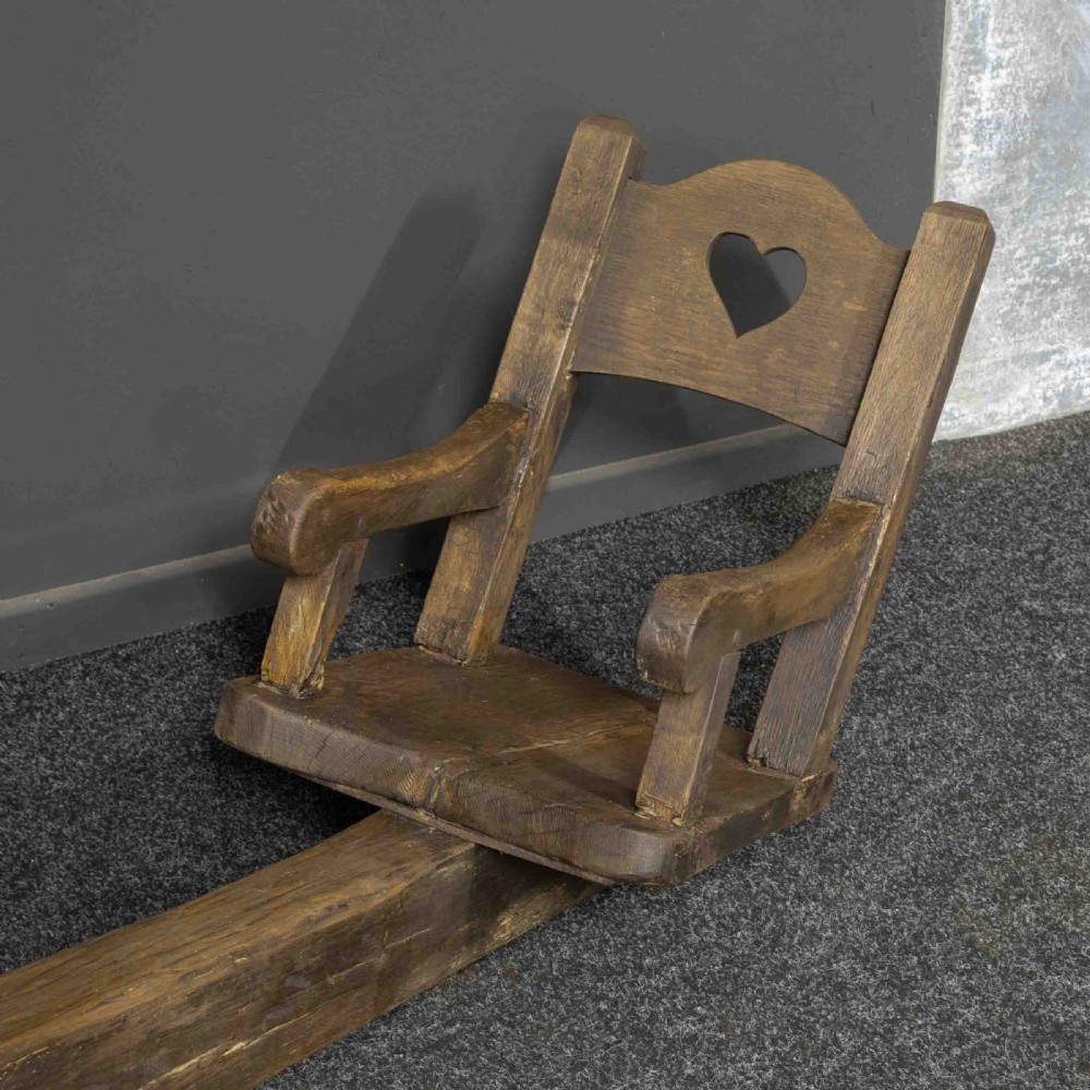 A large dark timber ducking stool featuring a long lever beam, a seated wooden chair at one end, and a heavy support frame on wooden wheels. View XVI