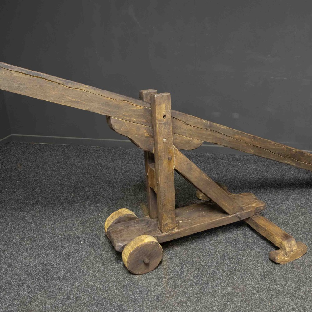 A large dark timber ducking stool featuring a long lever beam, a seated wooden chair at one end, and a heavy support frame on wooden wheels. View XVII