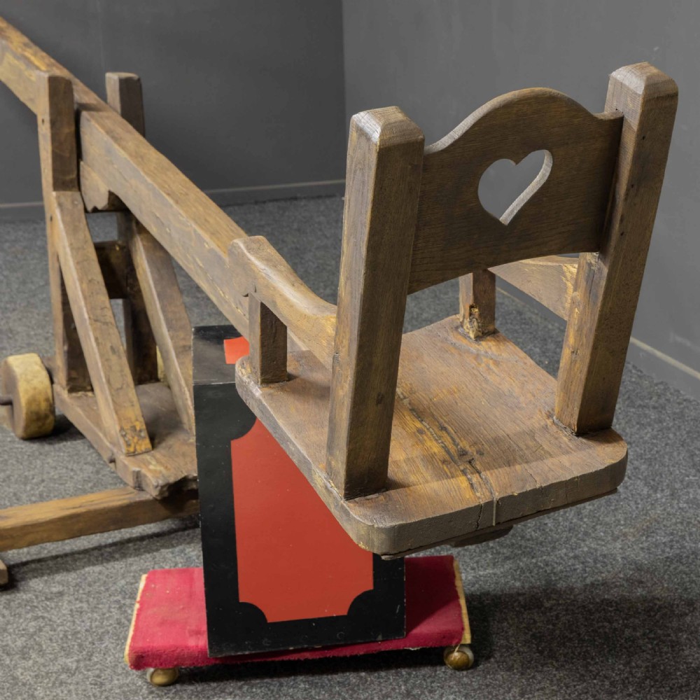 A large dark timber ducking stool featuring a long lever beam, a seated wooden chair at one end, and a heavy support frame on wooden wheels. View XVIII