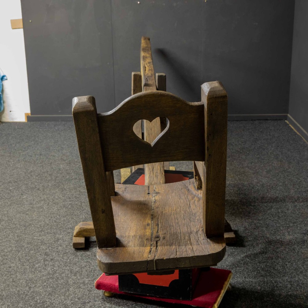 A large dark timber ducking stool featuring a long lever beam, a seated wooden chair at one end, and a heavy support frame on wooden wheels. View XIX