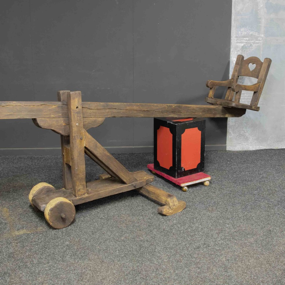 A large dark timber ducking stool featuring a long lever beam, a seated wooden chair at one end, and a heavy support frame on wooden wheels. View XX