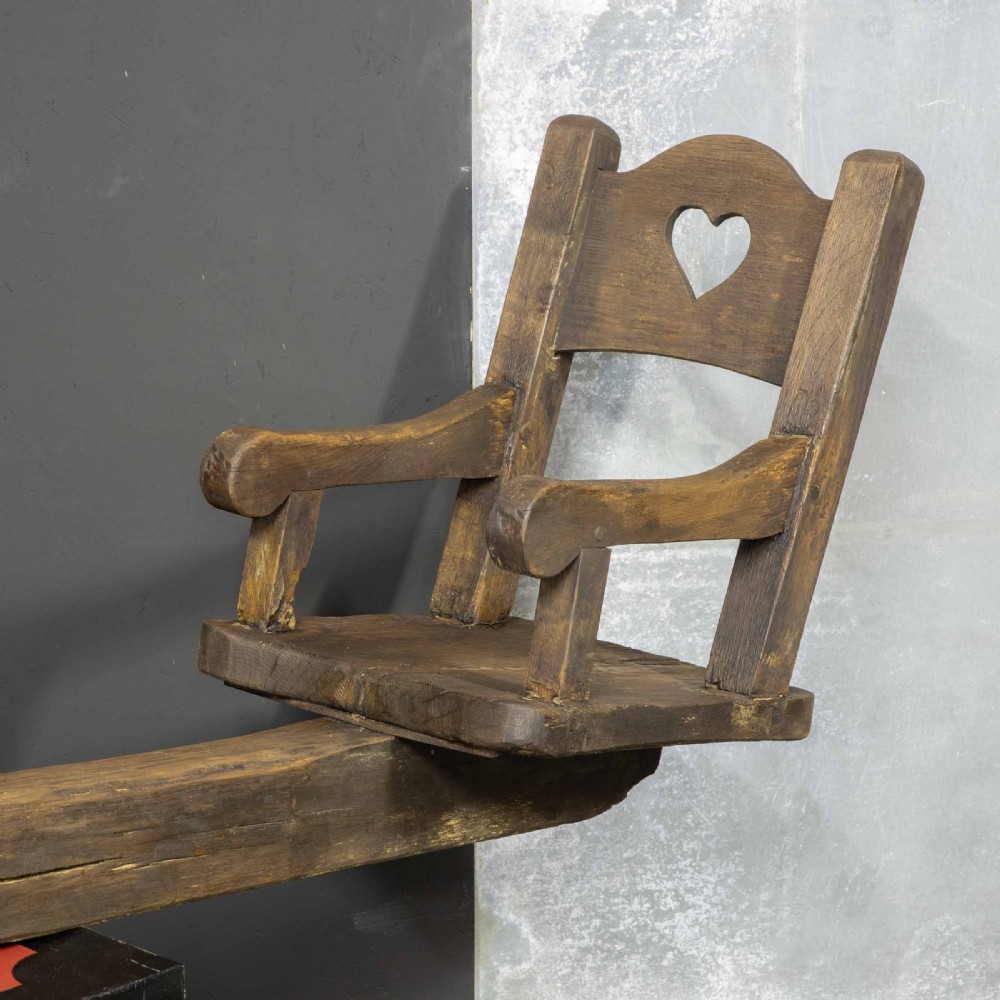 A large dark timber ducking stool featuring a long lever beam, a seated wooden chair at one end, and a heavy support frame on wooden wheels. View XXI