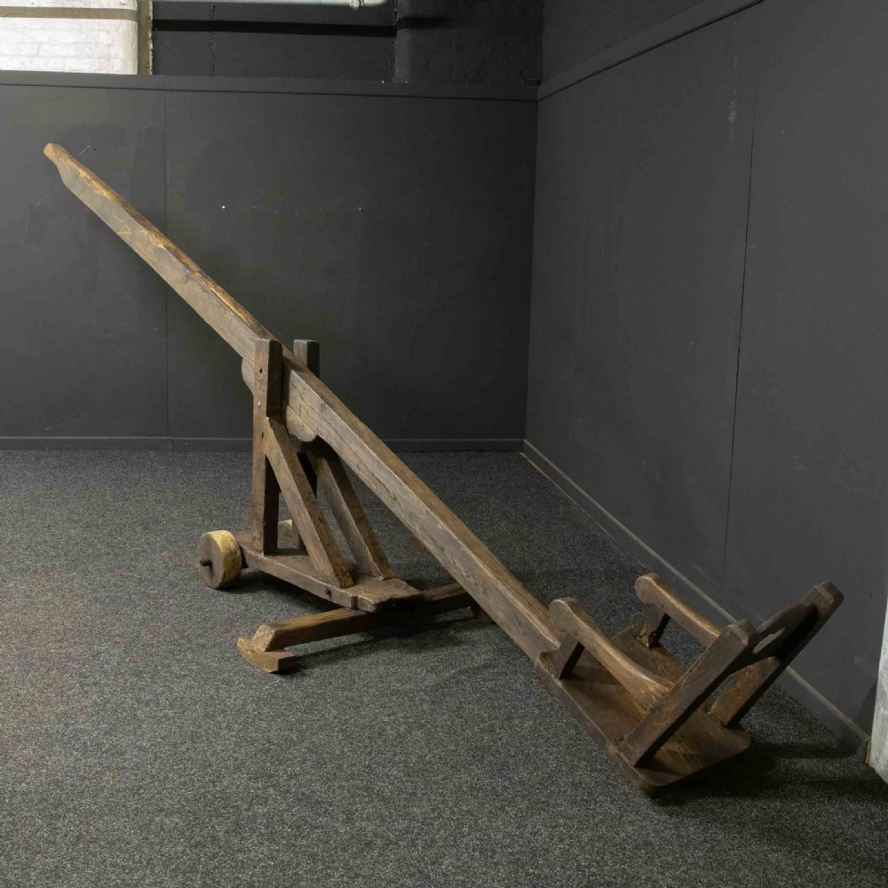 A large dark timber ducking stool featuring a long lever beam, a seated wooden chair at one end, and a heavy support frame on wooden wheels. View XXII