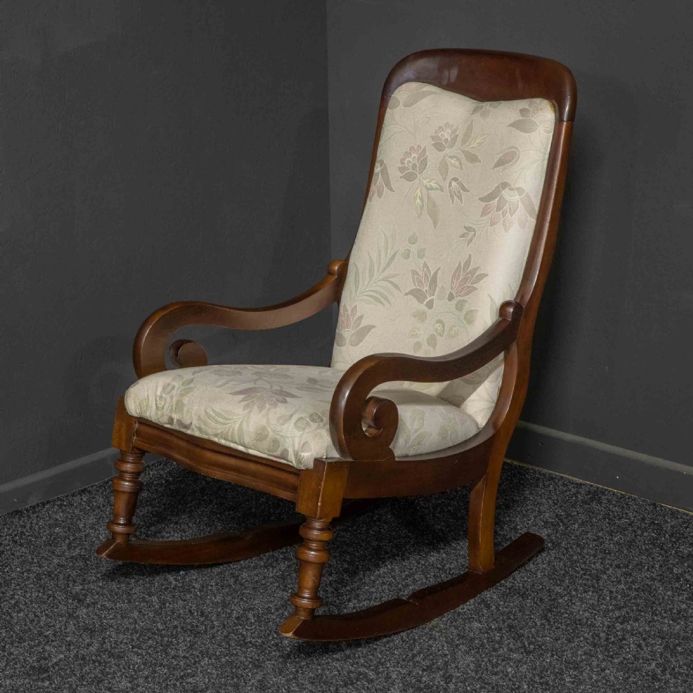 A Victorian rocking chair crafted from dark polished mahogany with scroll arms, turned front legs, and cream floral upholstery.
