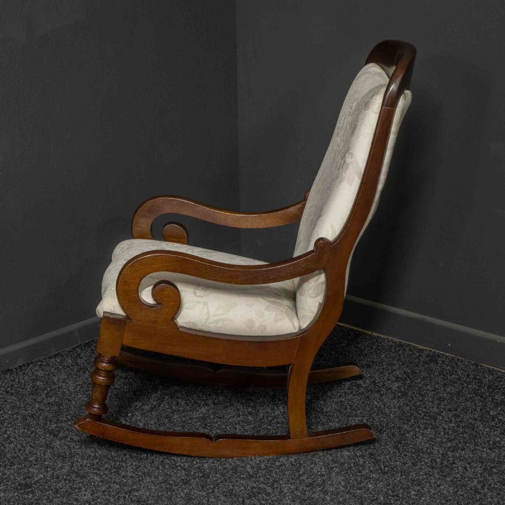 A Victorian rocking chair crafted from dark polished mahogany with scroll arms, turned front legs, and cream floral upholstery. View VI