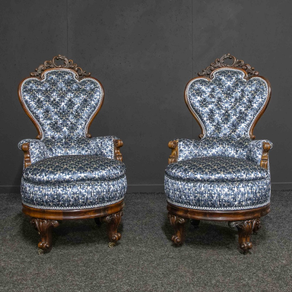 A pair of Victorian armchairs with carved timber frames, blue patterned button-back fabric upholstery, and rounded seat cushions on brass castors. View V
