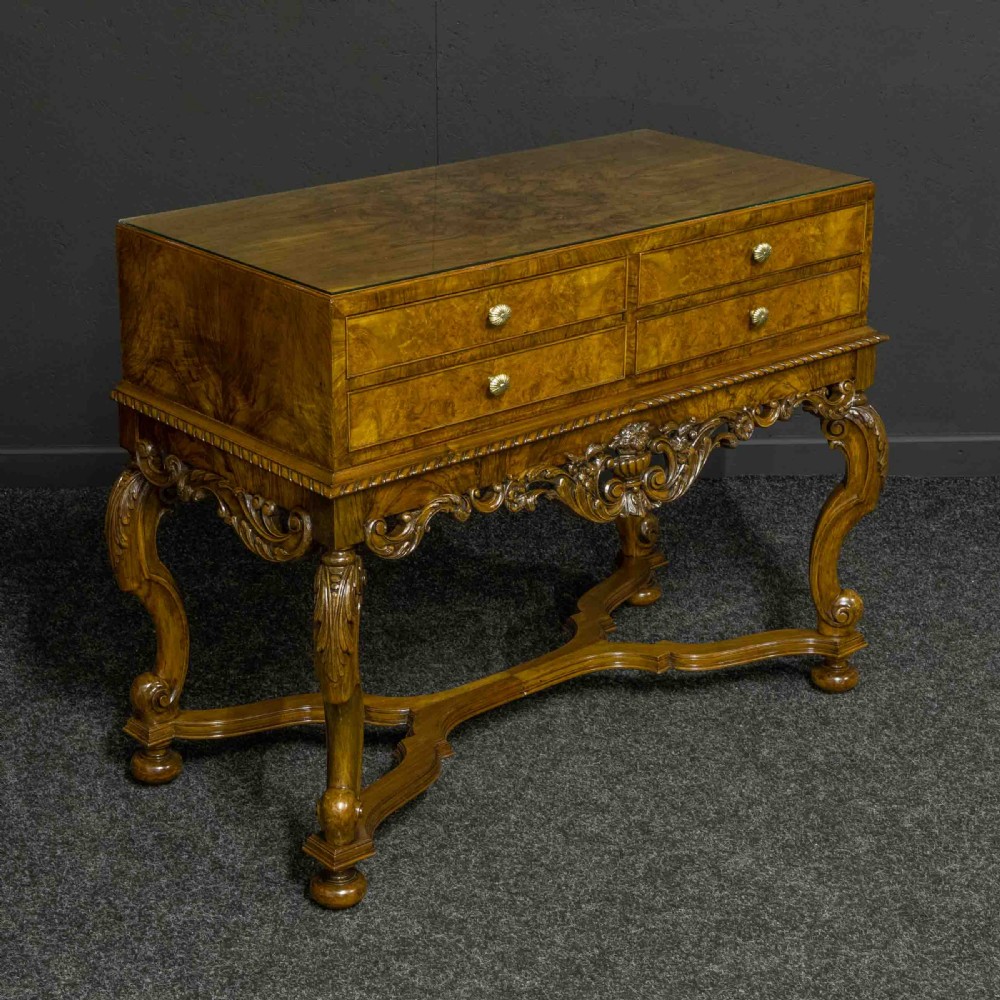 A burr walnut chest on stand featuring four drawers with brass knobs, an intricately carved foliate apron, cabriole legs, and a shaped stretcher base.
