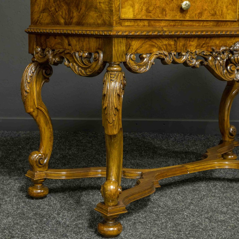 A burr walnut chest on stand featuring four drawers with brass knobs, an intricately carved foliate apron, cabriole legs, and a shaped stretcher base. View VII