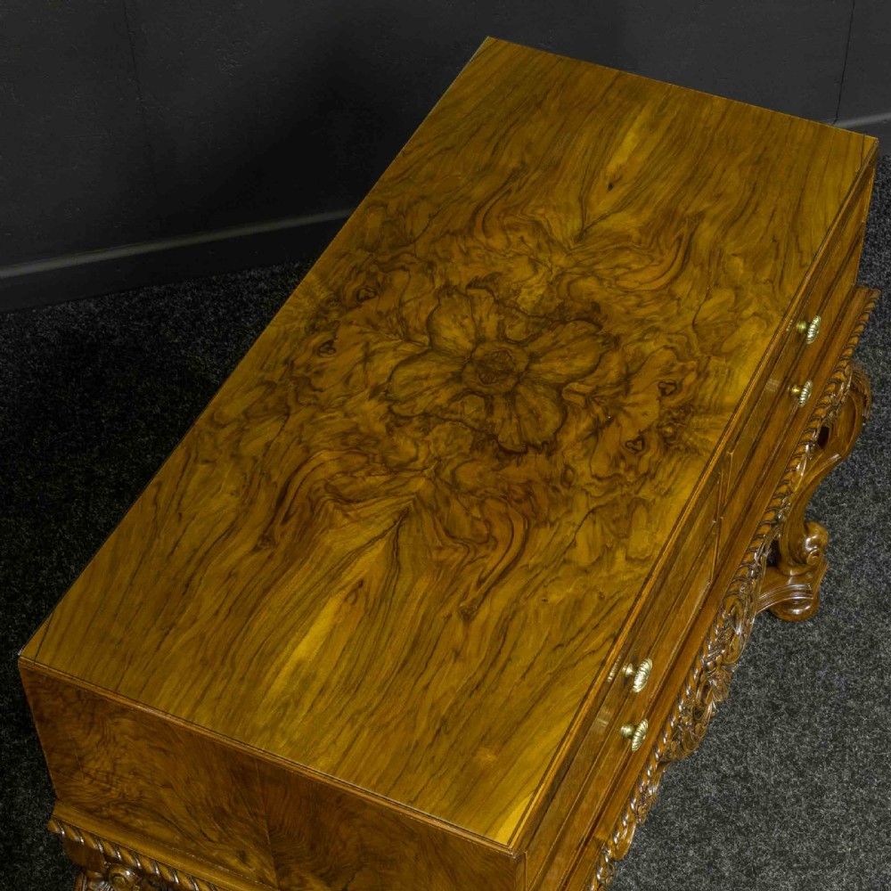 A burr walnut chest on stand featuring four drawers with brass knobs, an intricately carved foliate apron, cabriole legs, and a shaped stretcher base. View XIII
