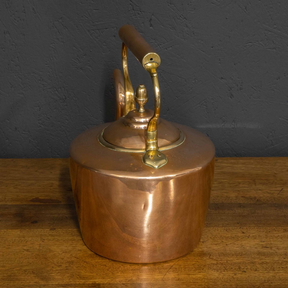 A polished Victorian copper kettle featuring a brass finial lid, a curved spout, and an overhead copper handle with brass supports. View V