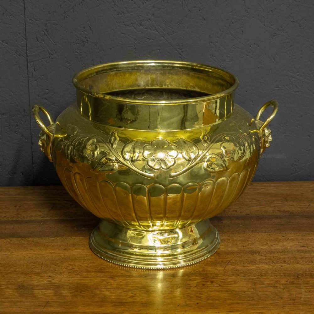 A polished brass planter featuring embossed floral motifs, gadrooned lower body detailing, twin side handles, and a raised pedestal base.