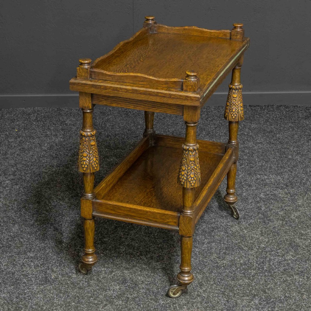A dark oak two-tier tea trolley with carved turned legs, a galleried top tray, and metal castors.