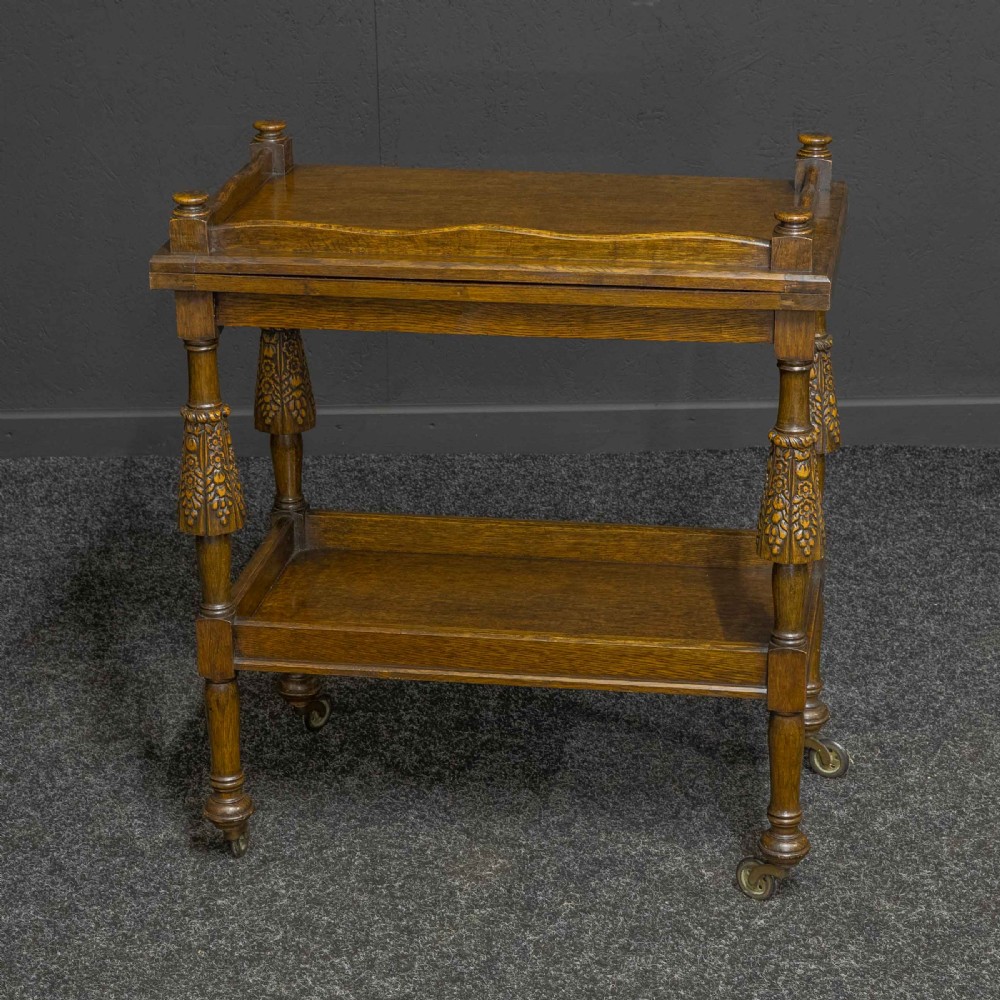 A dark oak two-tier tea trolley with carved turned legs, a galleried top tray, and metal castors. View III
