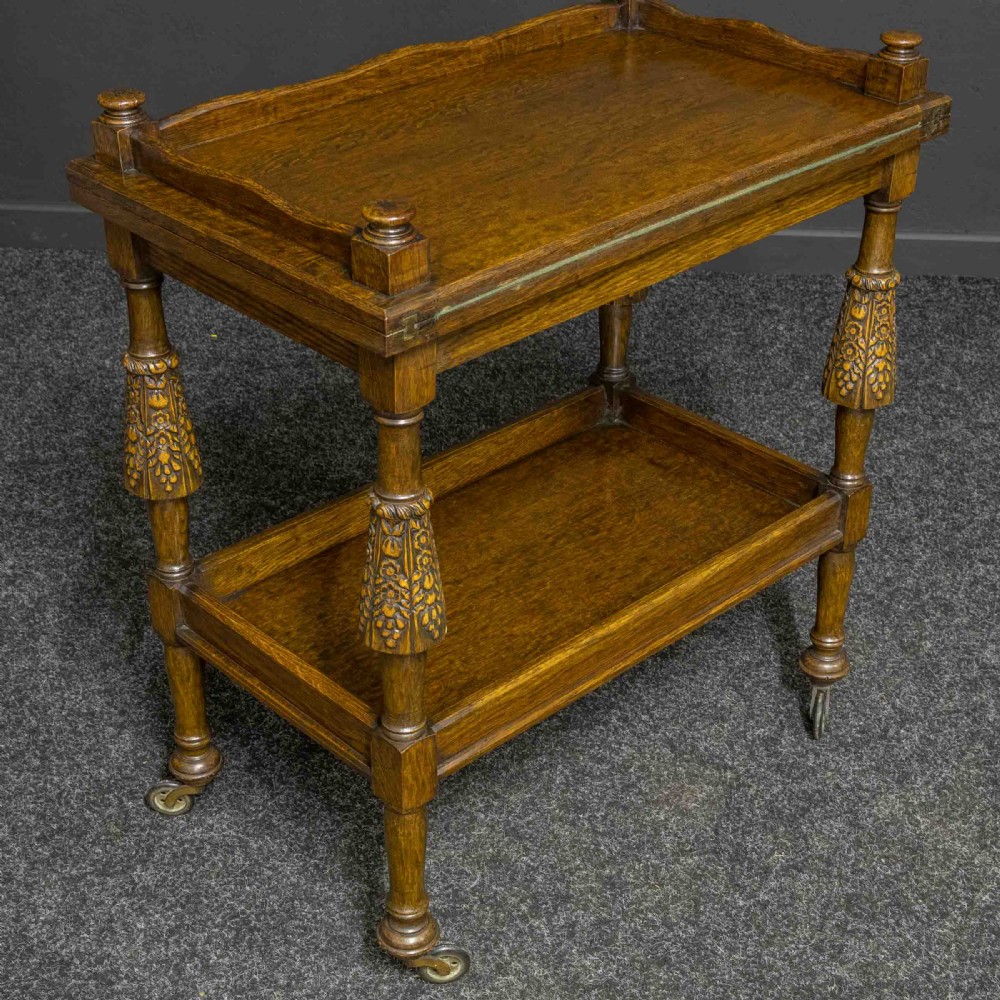 A dark oak two-tier tea trolley with carved turned legs, a galleried top tray, and metal castors. View VII