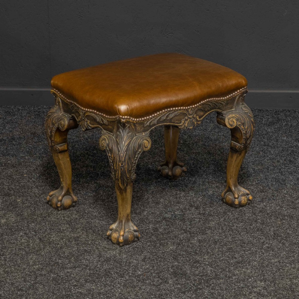 A low Chippendale style stool featuring a brown leather upholstered seat with brass stud detailing and dark carved cabriole legs ending in claw and ball feet.