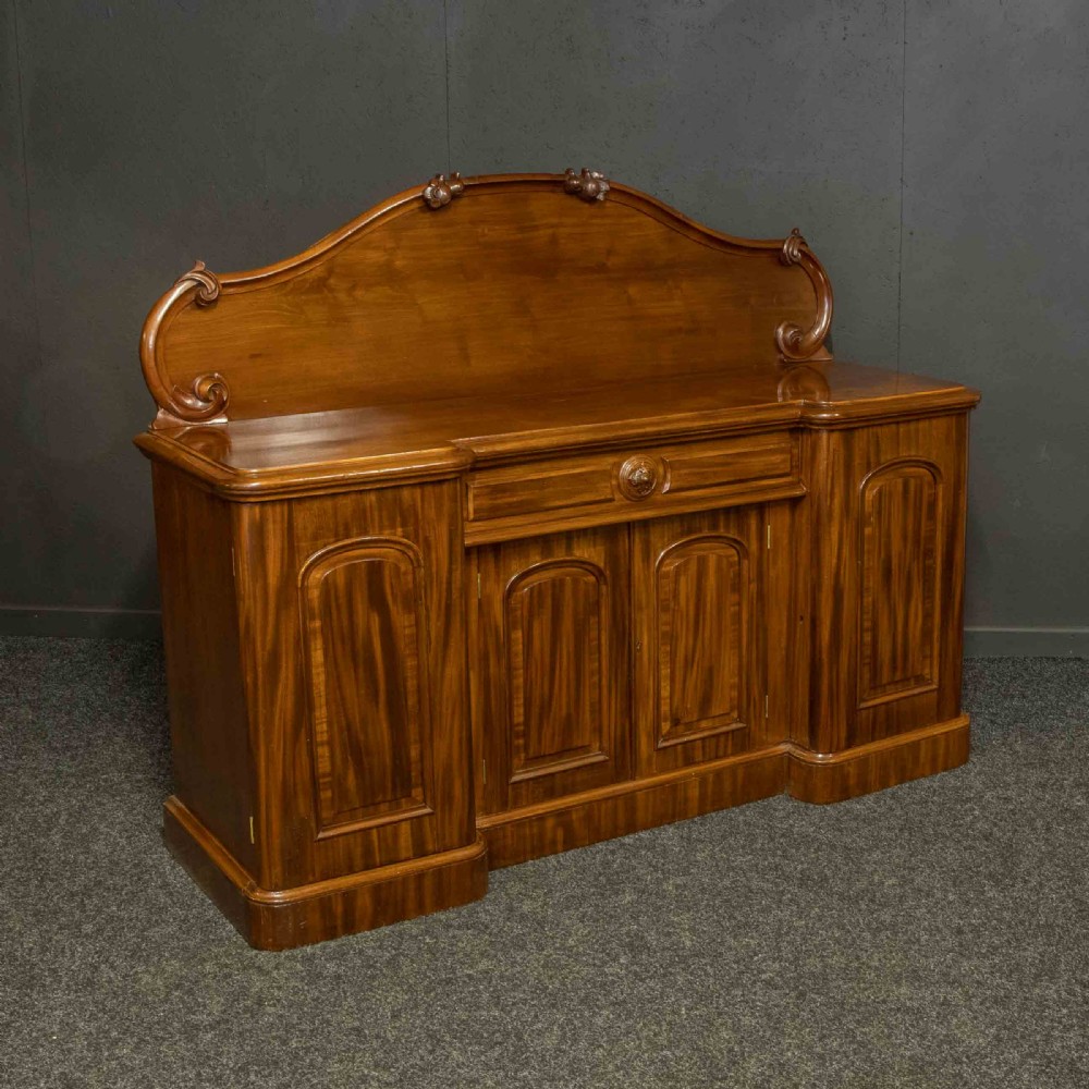 A Victorian mahogany sideboard featuring a carved backboard gallery, flame mahogany panelled doors, a central drawer, and a plinth base.
