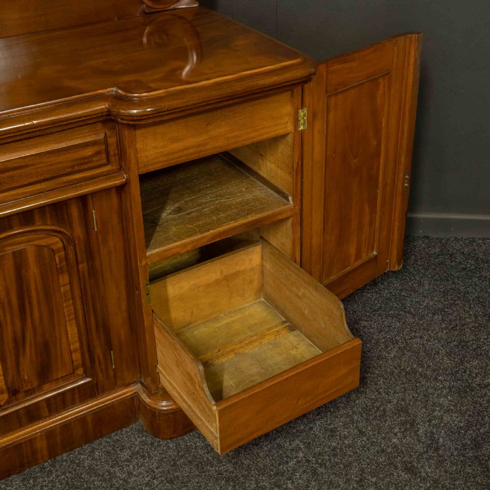 A Victorian mahogany sideboard featuring a carved backboard gallery, flame mahogany panelled doors, a central drawer, and a plinth base. View IV