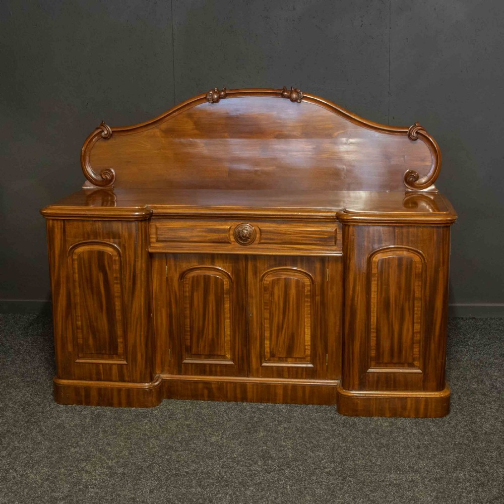 A Victorian mahogany sideboard featuring a carved backboard gallery, flame mahogany panelled doors, a central drawer, and a plinth base. View V