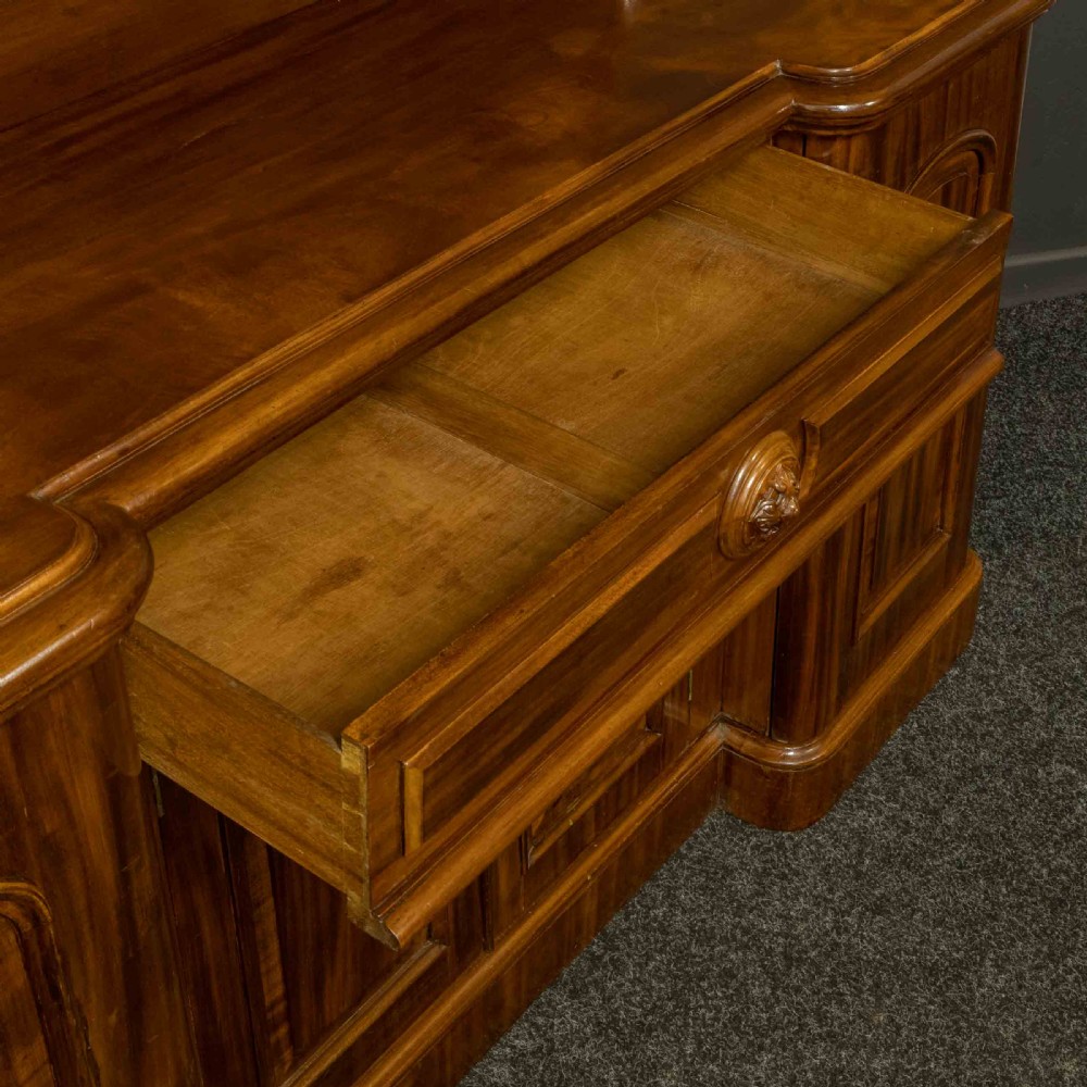 A Victorian mahogany sideboard featuring a carved backboard gallery, flame mahogany panelled doors, a central drawer, and a plinth base. View VI