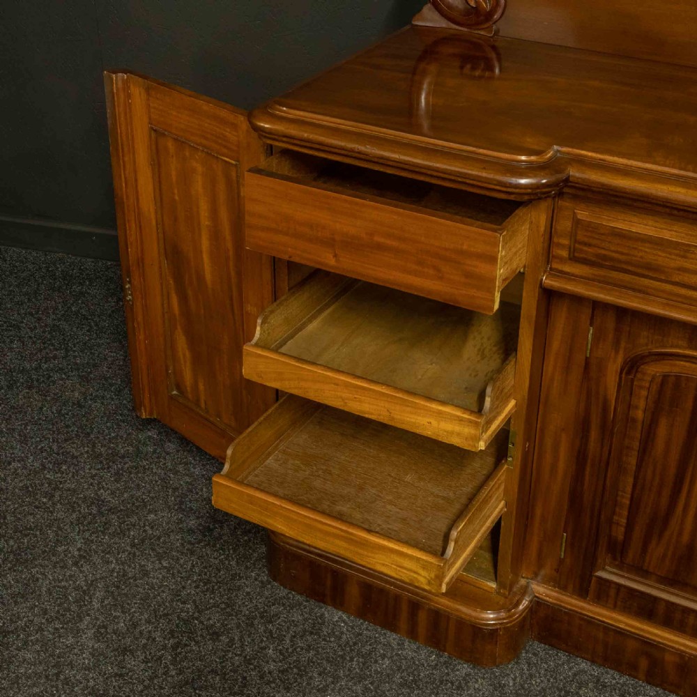 A Victorian mahogany sideboard featuring a carved backboard gallery, flame mahogany panelled doors, a central drawer, and a plinth base. View VIII