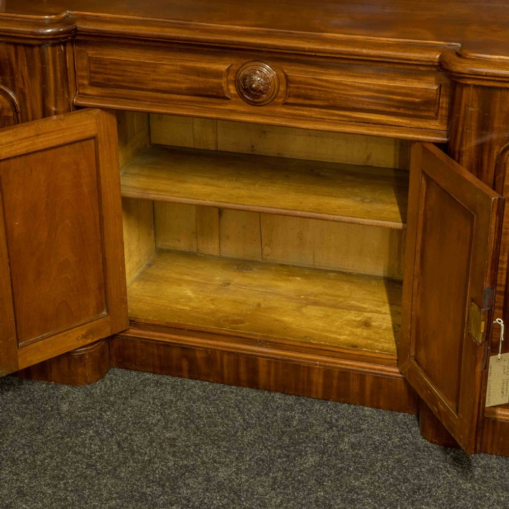A Victorian mahogany sideboard featuring a carved backboard gallery, flame mahogany panelled doors, a central drawer, and a plinth base. View XI