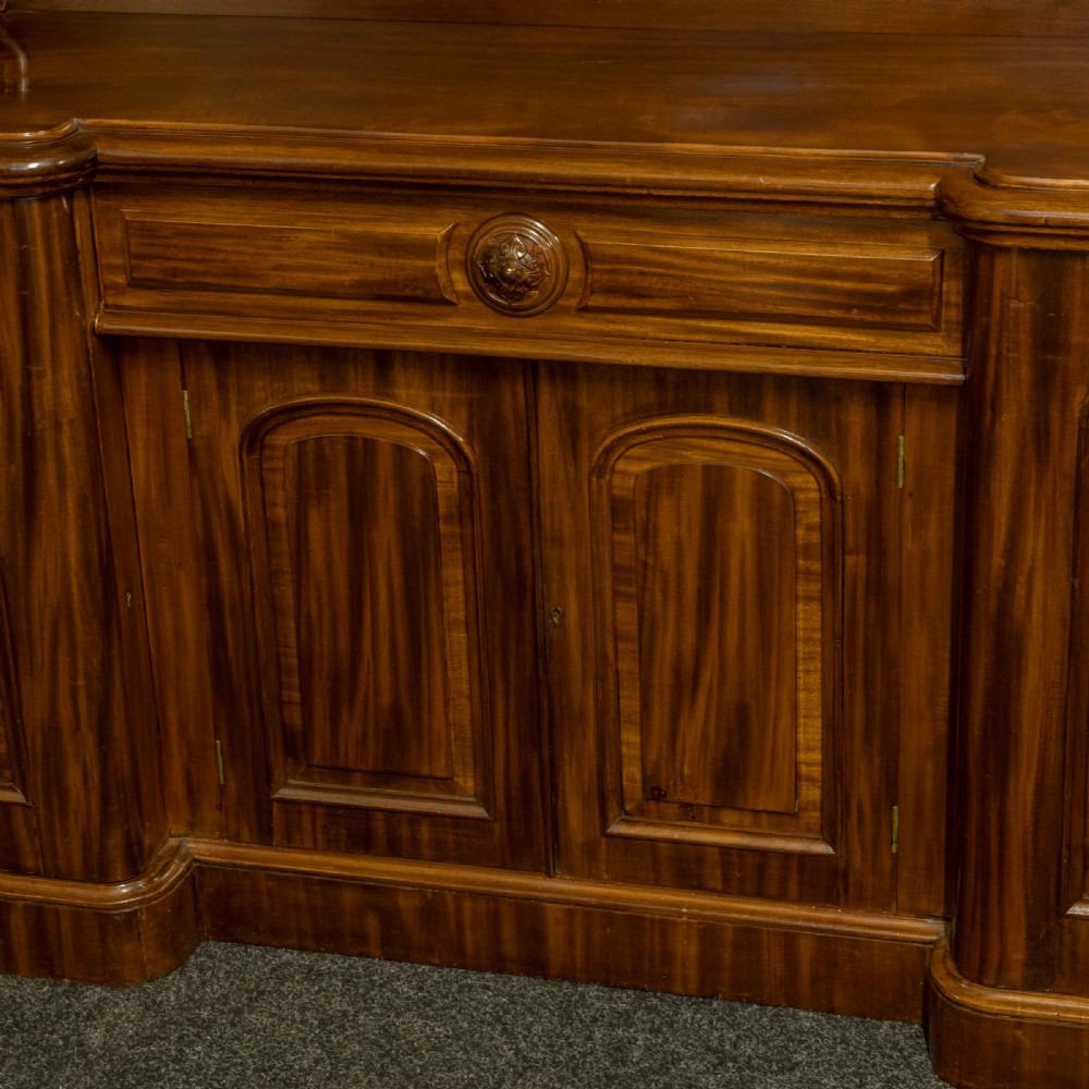 A Victorian mahogany sideboard featuring a carved backboard gallery, flame mahogany panelled doors, a central drawer, and a plinth base. View XII
