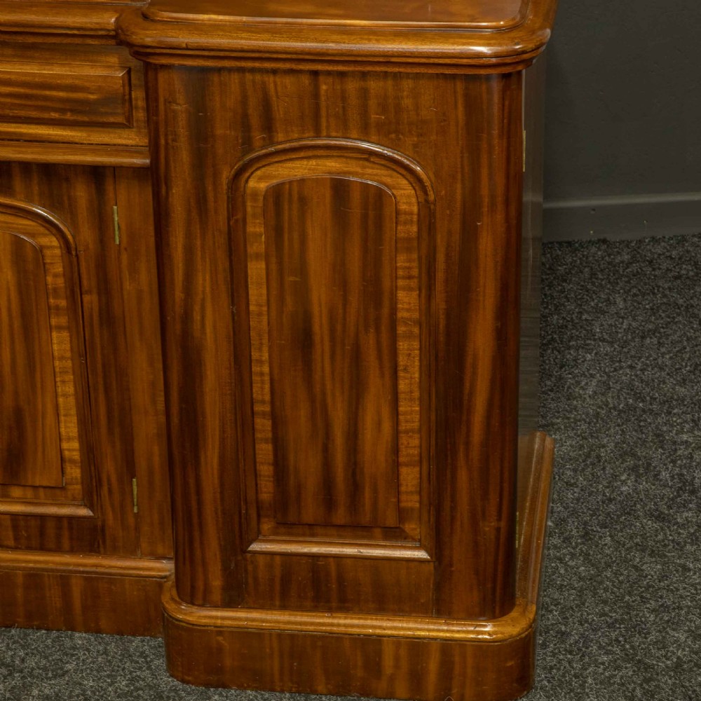 A Victorian mahogany sideboard featuring a carved backboard gallery, flame mahogany panelled doors, a central drawer, and a plinth base. View XIV