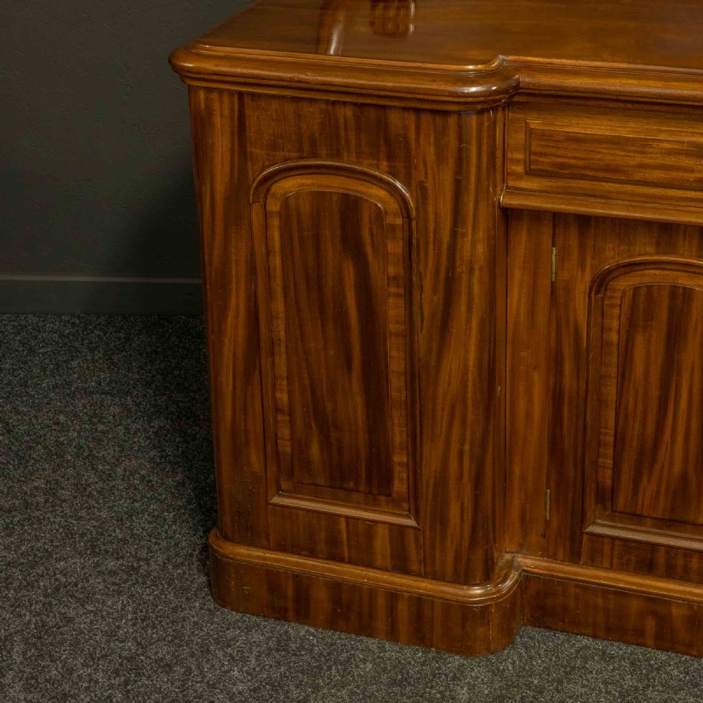 A Victorian mahogany sideboard featuring a carved backboard gallery, flame mahogany panelled doors, a central drawer, and a plinth base. View XV