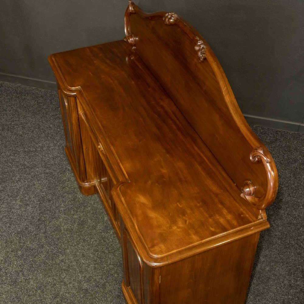 A Victorian mahogany sideboard featuring a carved backboard gallery, flame mahogany panelled doors, a central drawer, and a plinth base. View XVII