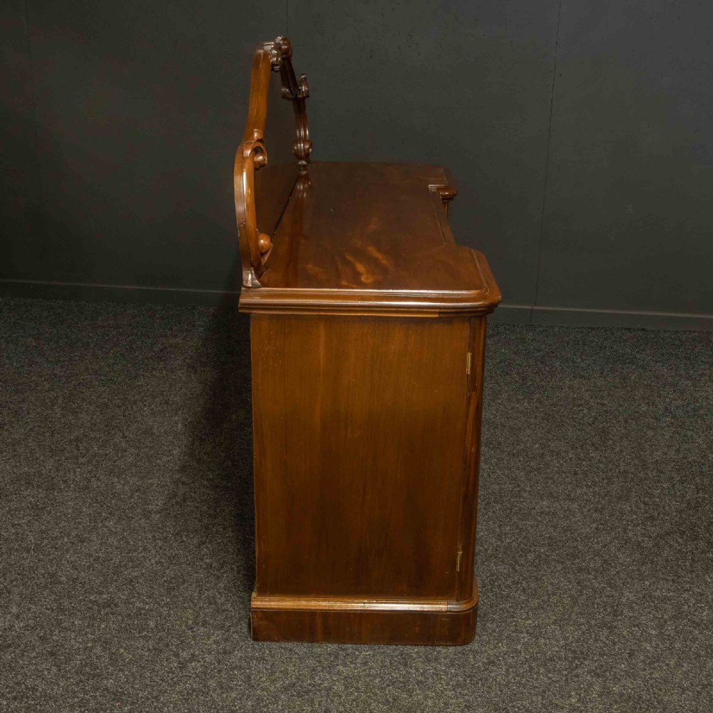 A Victorian mahogany sideboard featuring a carved backboard gallery, flame mahogany panelled doors, a central drawer, and a plinth base. View XX