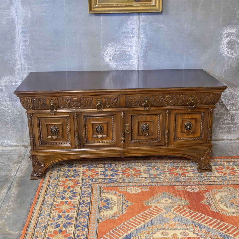 A low carved dark walnut sideboard featuring four panelled doors with cast brass lion mask drop handles below a carved relief frieze.