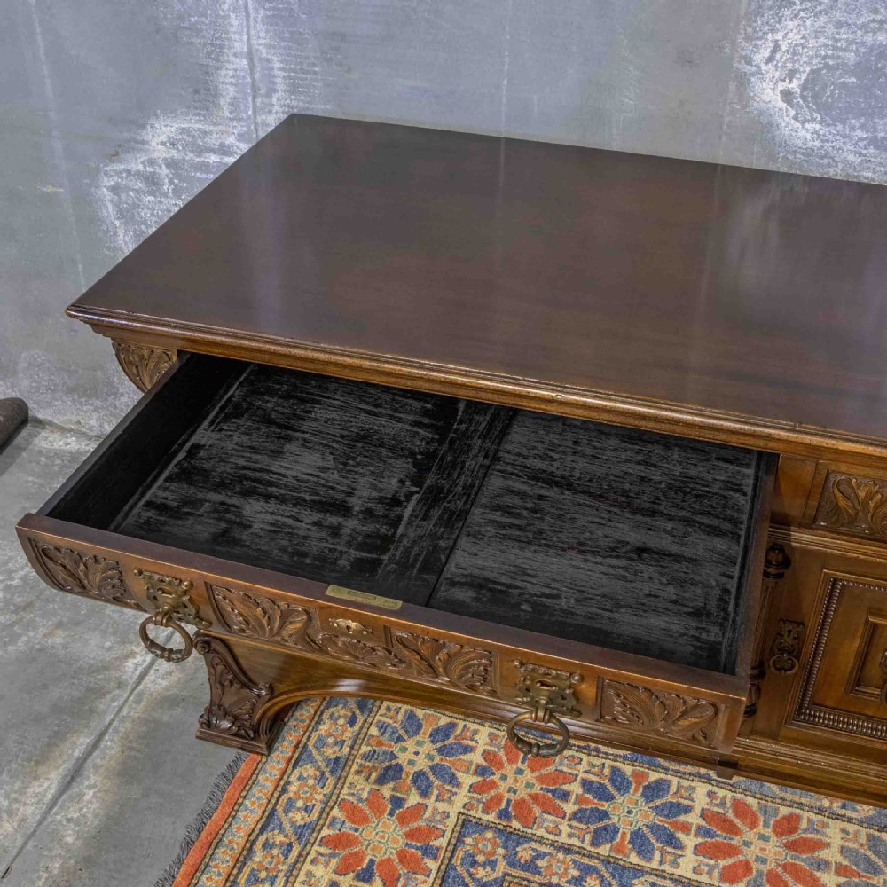 A low carved dark walnut sideboard featuring four panelled doors with cast brass lion mask drop handles below a carved relief frieze. View II