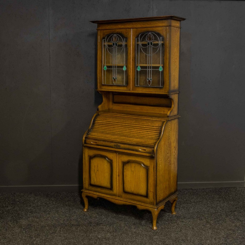 An Edwardian oak roll top bureau bookcase featuring an upper cabinet with two leaded glass doors, a central curved tambour writing section, and a two-door lower cupboard.