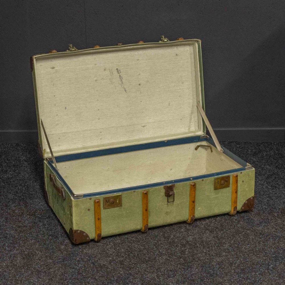 An olive green canvas-covered steamer trunk featuring wooden bentwood slats, leather corner protectors, leather side handles, and stencilled black lettering. View V