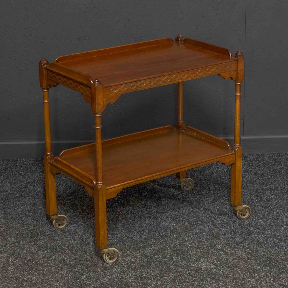 A two-tiered warm brown polished wooden tea trolley featuring a carved geometric frieze, turned column supports, gallery edges, and four small metal castors.