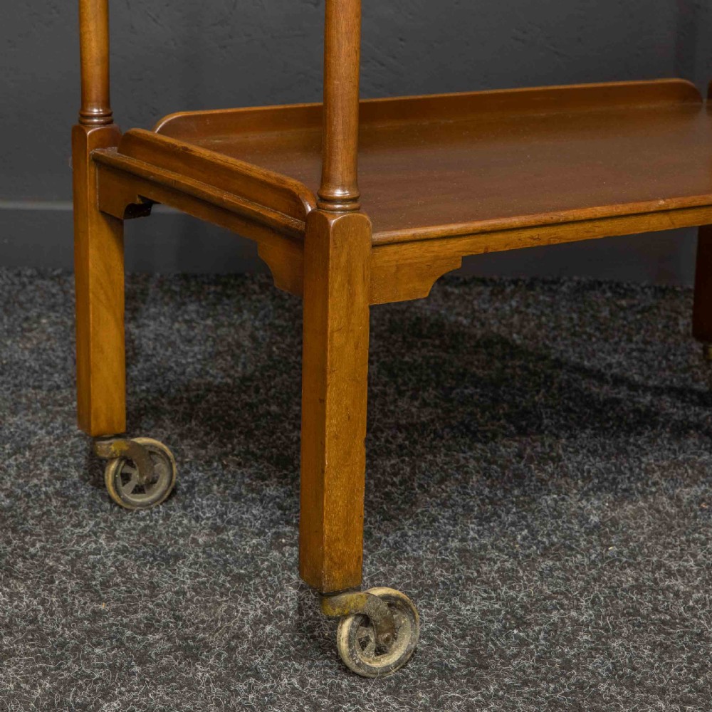 A two-tiered warm brown polished wooden tea trolley featuring a carved geometric frieze, turned column supports, gallery edges, and four small metal castors. View II